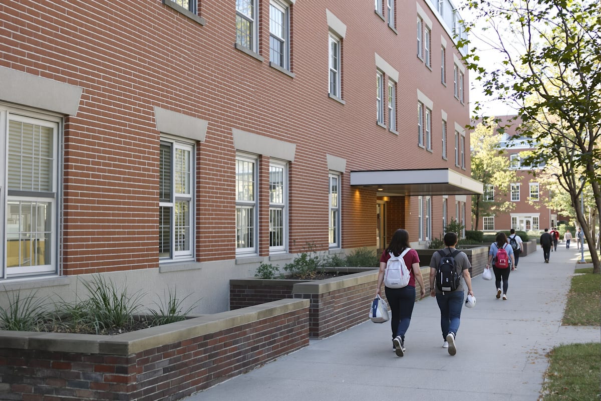 Students walking along a path beside a university residence hall in the morning.