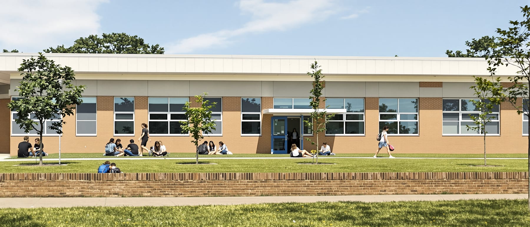 Students sitting and talking on a low wall outside a single-storey school on a sunny afternoon.